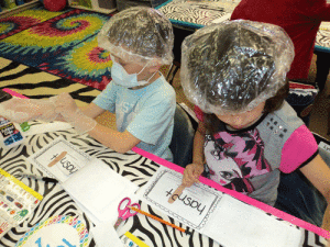 First graders in Mrs. Anderson’s room became surgeons for a day while learning how to turn 2 words into a contraction.  They dressed the part with surgical masks, hair nets and gloves. Next they “operated” by cutting out parts of words, removing letters and stitching them back together.  When they were finished, they put a bandaid over the scar (apostrophe)  creating a contraction. Picture Surgeons; Cole Spicak and Lyric Lopez operate on the words “has” and “not” to create the word hasn’t 